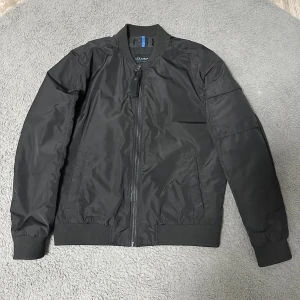 Svart bomberjacka från LCW Casual - Svart bomberjacka från LCW Casual i storlek L. Jackan har klassisk ribbad krage, muddar och nederkant, samt dragkedja framtill. Tillverkad i lätt och glansig polyester med fickor på sidorna och en extra ficka på ärmen. Perfekt för en chill och stilren look.