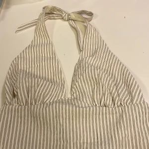 Helt ny randig maxiklänning i linnetyg med knyt i nacken - Naturvit, randig maxiklänning i linnetyg med halterneck med tunna bruna ränder och knyt i nacken och öppen rygg med knytband. Luftigt material, perfekt för varma dagar. Snygg v-ringning framtill och avslappnad passform.