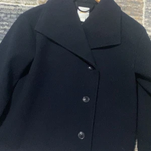 Svart kappa från Gina Tricot - Stilren svart kappa från Gina Tricot med bred krage och tre knappar framtill. Klassisk rak passform och långärmad design, perfekt för dig som gillar enkel och snygg look. Materialet är mjukt och känns skönt mot kroppen.