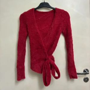 Röd fluffig omlottopp med knyt - Säljer en röd fluffig omlottopp med långa ärmar och v-ringad hals. Toppen har ett snyggt knyt i midjan och är gjord i ett mjukt, lurvigt material som känns supermysigt. Perfekt för dig som gillar statement-plagg med cool textur och färg.