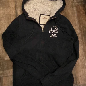 Fodrad Marinblå zip hoodie från Hollister  - Mysig marinblå hoodie från Hollister med fluffigt vitt foder i huvan. Tröjan har dragkedja, snörning vid huvan och broderad logga på bröstet. Perfekt för kyliga dagar och har praktiska fickor framtill.