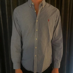 Rutig blå skjorta från Ralph Lauren - Snygg rutig skjorta från Ralph Lauren i blått och vitt med klassisk button-down krage och långärmad passform. Skjortan har knappar framtill och en liten röd logga broderad på bröstet. Perfekt för dig som gillar stilrena och tidlösa plagg.