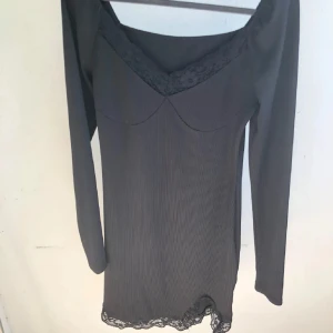 Svart långärmad klänning med spets - Snygg svart kortklänning med långa ärmar och v-ringad halsringning dekorerad med spets. Klänningen har en figurnära passform och ribbad struktur, samt spetsdetaljer vid nederkanten, mycket stretch i den är S men passar M oxå