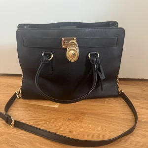 Svart handväska från Michael Kors - Stilren svart handväska från Michael Kors i skinn med guldfärgade metalldetaljer och kedja på axelremmen. Väskan har både handtag och justerbar axelrem samt ett snyggt MK-lås framtill. Perfekt för dig som gillar klassisk och lyxig design.