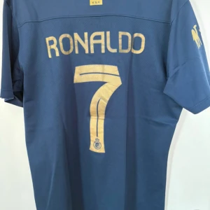 Al Nassr Ronaldo Nike fotbollströja L - Säljer en blå Al Nassr fotbollströja från Nike med guldiga detaljer, Ronaldo 7 på ryggen och klubbmärke framtill. Tröjan är i Dri-Fit material och har korta ärmar. Perfekt för dig som vill bära samma stil som Ronaldo på planen.