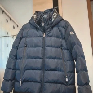 Mörkblå Moncler pufferjacka L - Säljer en mörkblå pufferjacka från Moncler Genius med stora vita logotexter på huvan och detaljerade dragkedjor. Jackan har två sidofickor med dragkedja och en quiltad design som ger en schysst volym. Perfekt för kalla dagar och riktigt snygg streetstil.