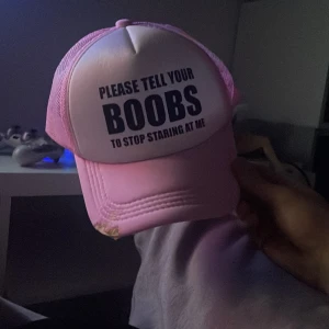 Rosa och vit truckerkeps med text - Säljer en rosa och vit truckerkeps med svart tryck på framsidan där det står 'PLEASE TELL YOUR BOOBS TO STOP STARING AT ME'. Kepsen har mesh på sidorna och bak, samt böjd skärm. Perfekt för dig som gillar statement-plagg och vill sticka ut.