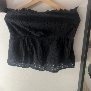Svart ärmlös tubtopp med spetsdetaljer - Säljer en svart tubtopp med broderade hål och spetskant upptill och nedtill. Toppen är axelbandslös och har en volangliknande nederdel som ger en söt och luftig känsla. Perfekt för dig som gillar en feminin och trendig stil.