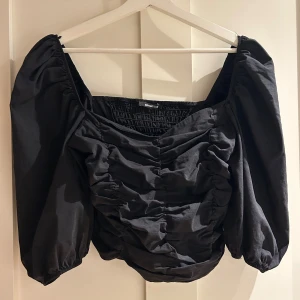 Svart puffärmad blus från Gina Tricot - Supersnygg svart blus från Gina Tricot med puffiga långa ärmar och smockad rygg. Framsidan har trendiga rynkade detaljer och en lätt v-ringad hals. Perfekt för dig som vill sticka ut med en cool och modern look.