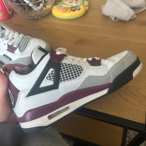 Nike Jordan 4 Bordeaux sneakers - Nike Jordan 4 Bordeaux sneakers med vit ovandel i skinn, detaljer i vinrött och grått, svarta inslag och meshpaneler på sidorna. Klassisk Jumpman-logga på plösen och snörning framtill. Snygg och ikonisk siluett med platt sula och rund tå.PSG edition