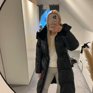 Svart lång pufferjacka med huva - Säljer en svart, lång pufferjacka med stor huva och dragkedja framtill. Jackan har quiltad design, snörning i midjan och en cool patch på ärmen. Perfekt för kalla dagar 