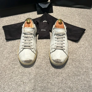 Saint Laurent vita sneakers med svart häl - Säljer ett par Saint Laurent sneakers i vitt skinn med handskriven logga på sidan och svart detalj vid hälen. Skorna har rund tå, platt sula och snörning. Skorna är relativt slitna i skiner på vissa ställen, men är fortfarande helt fungerade utan några skador mer en dom estetiska skadorna på tårna fram. Allting som kom med när jag köpte skorna finns. Kvitto, extra sko snören, lådan och påsen. Skorna är köpte i Saint Lorant butiken i New York för 775 dollar.