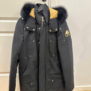 Svart parkajacka från Moose Knuckles Stirling Stolek M - Svart parkajacka från Moose Knuckles med guldfärgade detaljer och dragkedja. Jackan har en stor huva med svart päls och flera fickor framtill. Insidan har ett broderat motiv med hockeyspelare och älg. Perfekt för kalla dagar och riktigt snygg stil. Använd 2 tillfälle  förra vintern inprincip helt ny! 