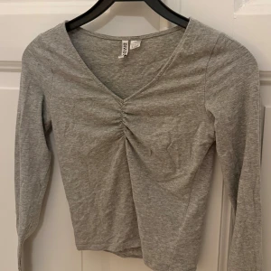 Grå långärmad topp - Säljer en grå långärmad topp från H&M DIVIDED i storlek S. Toppen har en snygg v-ringning med rynkade detaljer framtill och är tillverkad i mjuk bomull. Endast använd några fåtal gånger!