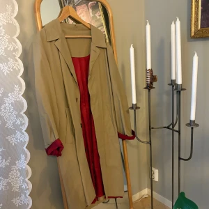 Beige trenchcoat med röd insida - Säljer en klassisk beige trenchcoat med snygg röd insida. Jackan har krage, knappar framtill och två fickor på sidorna. Perfekt för dig som gillar stilrena och tidlösa plagg med en twist. Passar dig som vill ha en längre jacka med cool färgdetalj. Jag skulle säga att den är i storlek one size.