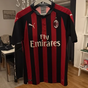 AC Milan fotbollströja 2018/2019 (Äkta med Serie A och CL Logo) - AC Milan matchtröja från säsongen 2018/19 med klassiska röda och svarta vertikala ränder, korta ärmar och broderade Puma-loggor. Materialet är lätt och ventilerande polyester. Serie A och Champions League-märken på ärmarna samt Fly Emirates tryck på bröstet.