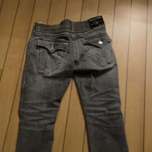 True Religion Ricky grå jeans straight - Säljer ett par grå jeans från True Religion, modell Ricky Relaxed Straight. De har klassiska kontrastsömmar, raka ben och snygga fickdetaljer med knappar bak. Märkespatch på linningen och slitningar för en cool look. Dem är använda 1-6 gånger köptes ifrån jeansbolaget!