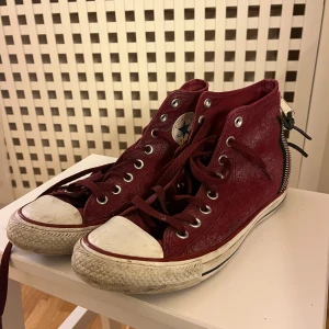 Vinröda Converse All Star high tops - Säljer ett par vinröda Converse All Star high tops med klassisk vit tå och sula. Skorna har snörning framtill, dragkedja bak och är tillverkade i textil med gummisula. Kedjor där bak.