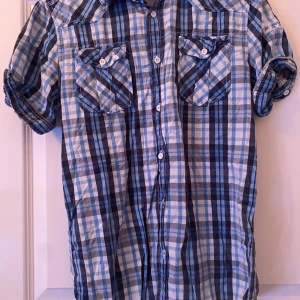 Blårutig kortärmad skjorta Blend  - Snygg blårutig skjorta från Blend i barn storlek XL. Skjortan har korta ärmar, två bröstfickor med knappar och klassisk krage. Perfekt för dig som gillar en avslappnad och cool stil. Tillverkad i mjuk bomull för skön känsla.