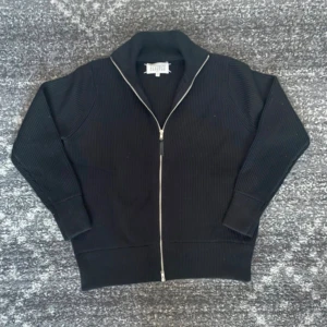 Svart  kofta från Maison Margiela - Svart ribbstickad kofta från Maison Margiela med hel dragkedja framtill och hög krage. Tröjan har långa ärmar och klassiska vita stygn i nacken. Perfekt för dig som gillar minimalistisk och stilren design.