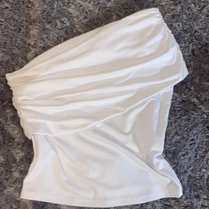 Vit oneshoulder topp från H&M Divided - Säljer en vit oneshoulder topp från H&M Divided i storlek S. Toppen har snygga draperade detaljer och är axelbandslös på ena sidan. Materialet är mjukt och stretchigt, perfekt för att skapa en cool och trendig look. Passar dig som gillar stilrena och moderna plagg.