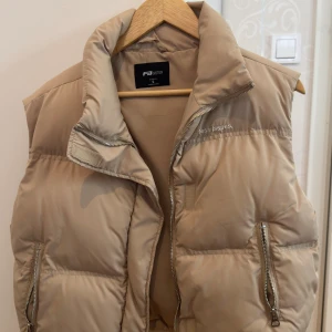 Beige dunväst från FB Sister - Snygg beige dunväst från FST Sister med hög krage, två stora fickor med dragkedja och broderad text 'less issues.' på bröstet. Västen har justerbar dragsko nertill och stängs med dragkedja och kardborre. Perfekt för lager-på-lager-stil.