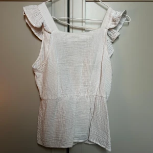 Vit blus med volang från Vero Moda - Supersöt vit blus från Vero Moda i storlek XS. Blusen har volangdetaljer vid axlarna och omlottöppning i ryggen. Tillverkad i mjuk bomull med lätt struktur, perfekt för sommarens alla tillfällen. Snygg att styla med jeans eller kjol! 