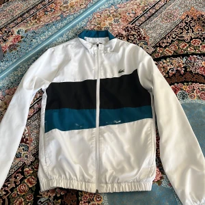 Lacoste Sport vindjacka XS - Snygg vindjacka från Lacoste Sport i vitt med breda svarta och blåa blockränder över bröstet. Jackan har dragkedja framtill, elastiska muddar och klassisk Lacoste-logga på bröstet. Perfekt för dig som gillar sportig och clean stil. Den har lite färger på sig annars den är äkta lacoste.
