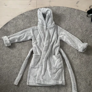 Grå Morgonrock med huva från Next - Supermjuk och fluffig grå morgonrock från Next med huva och stora fickor framtill. Morgonrocken har bälte i midjan och långa ärmar med uppvikta muddar. Perfekt för att mysa runt hemma eller när du vill ha något riktigt cozy på dig.