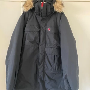  Fjällräven jacka  - Svart parkasjacka från Fjällräven med avtagbar huva och snygg fuskpäls. Jackan har flera stora fickor framtill, dragkedja och tryckknappar. Klassisk röd Fjällräven-logo på bröstet och detaljer i syntetmaterial. Perfekt för kalla vinterdagar. Säljer min morfars jacka, använd endast 1 gång i 1 timme 