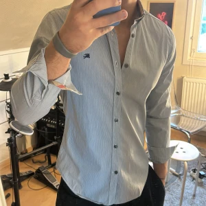 Randig skjorta från Burberry Brit - Snygg ljusblå randig skjorta från Burberry Brit med klassisk krage och svarta knappar. Långärmad modell med diskret Burberry-logga på bröstet och ikoniskt rutigt mönster på insidan av kragen. Tillverkad i mjuk bomull för skön känsla.