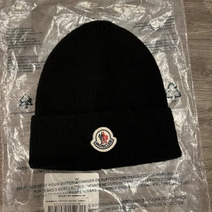 Svart Moncler mössa i ull - Snygg svart mössa från Moncler med klassisk ribbstickad design och uppvikt kant. Mössan har Moncler-logga framtill i vitt, rött och blått. Tillverkad i mjuk ull som håller dig varm och stilren under kyliga dagar.