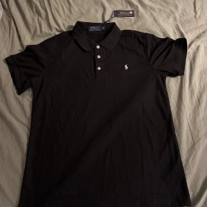 Svart pikétröja från Ralph Lauren - Klassisk svart pikétröja från Polo Ralph Lauren med vit broderad logga på bröstet. Tröjan har korta ärmar, tre knappar framtill och mjuk krage. Tillverkad i bomull för en skön och stilren känsla. Perfekt för dig som gillar enkel och tidlös stil.