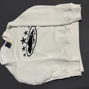 Vit långärmad tröja från Anyware - Snygg vit långärmad tröja från Anyware med svart tryck framtill som visar stjärnor och ett ovalt motiv. Tröjan har ribbade muddar vid ärmslut och nederkant, samt rund hals. Materialet är mjukt och känns som bomull, perfekt för chill dagar.