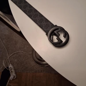 Svart Gucci GG Supreme bälte - Säljer ett svart Gucci GG Supreme bälte med klassisk GG-logga i matt svart metall. Bältet har ett diskret mönster med Guccis ikoniska GG-print och är tillverkat i skinn med textil på utsidan. Snyggt och stilrent för dig som gillar designeraccessoarer.