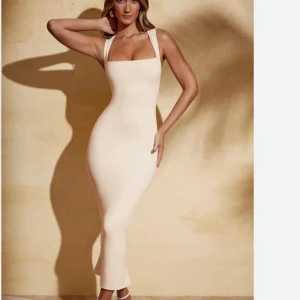 Champagnefärgad långklänning med breda axelband - Supersnygg långklänning i champagnefärg med breda axelband och fyrkantig urringning. Klänningen är figurnära och har en stilren, minimalistisk look som verkligen framhäver siluetten. Perfekt för dig som gillar en elegant och modern vibe.