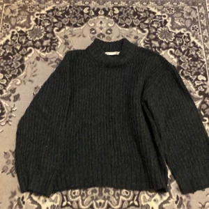 Mörkgrå stickad polotröja 157 - Mysig mörkgrå stickad polotröja från 157 med hög krage och ribbad struktur. Perfekt för kyliga dagar och enkel att matcha med jeans eller kjol. Tröjan har lång ärm och en loose fit som ger en avslappnad vibe.