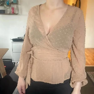 Beige omlottblus med prickar - Säljer en beige omlottblus med små prickar och knyt i midjan. Blusen har långärmade, lätt puffiga ärmar och v-ringad hals. Materialet är tunt och transparent, vilket ger en luftig känsla. Perfekt för dig som gillar detaljer och en feminin stil.