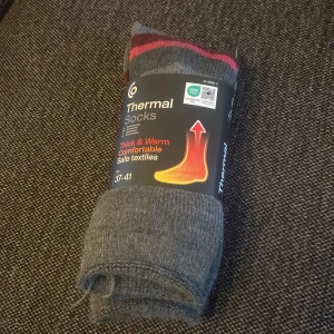 Grå termostrumpor - Säljer ett par grå termostrumpor från Clas Ohlson med diskreta röda och rosa ränder upptill. Strumporna är tjocka och värmande, perfekta för kalla dagar. Materialet är en blandning av bomull, polyester och polyamid för extra komfort och hållbarhet.