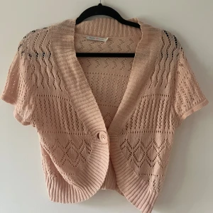 Rosa stickad kofta från ONLY - Säljer en rosa stickad kofta från ONLY med korta ärmar och ett snyggt hålmönster. Koftan har en bred ribbad krage och stängs med en stor knapp framtill. Perfekt att slänga över en topp för en chill look. Den är i storlek L men passar mer som S/M.