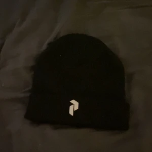 Svart peak mössa - Svart mössa med uppvikt kant och en stilren vit logga framtill. Enkel och clean design som passar till de flesta outfits. Perfekt för kyliga dagar när du vill hålla dig varm och samtidigt se snygg ut.