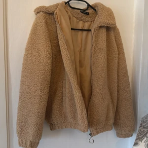 Beige teddyjacka från Ambar - Mysig teddyjacka i beige från Ambar, storlek S. Jackan har en fluffig och mjuk yta, dragkedja med rund metallring, krage och långa ärmar. Perfekt för kalla dagar och har en loose passform med elastisk nederkant och ärmslut.