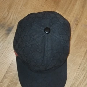 Svart Gucci keps med GG-mönster - Snygg svart keps från Gucci med klassiskt GG-mönster över hela kepsen. På sidan finns en grön och röd rand som ger extra lyxig känsla. Kepsen har böjd skärm och är tillverkad i bomull. Perfekt för dig som vill ha en stilren och exklusiv look. Äkta såklart lite solbränd men det märks inte 