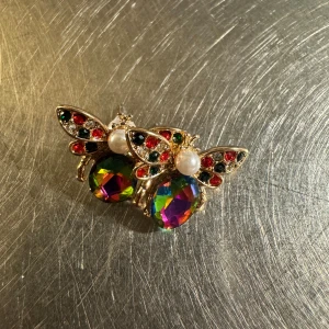Färgglada bi-örhängen med pärlor i 18k guldpläterad  - Unika örhängen i form av bin med 18k guldpläterad , vingar dekorerade med röda, svarta och vita stenar samt en pärla som huvud. Kroppen är en stor multifärgad kristall som skiftar i lila, grön och gul. Perfekt för dig som gillar statement-smycken och färg!