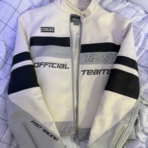 Race to Win skinnjacka i vitt och grått - Snygg skinnjacka från FB sister med coola racing-inspirerade detaljer och broderade texter som 'Official Team' och 'Fast Racing'. Jackan är vit med grå och svarta paneler, har dragkedja framtill och på ärmarna. Perfekt för dig som gillar motorsport och streetstyle.