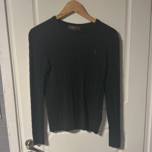 Mörkgrön stickad tröja från Morris - Snygg mörkgrön stickad tröja från Morris i 100% merinoull. Tröjan har ett klassiskt kabelstickat mönster, rund hals och lång ärm. På bröstet finns en broderad logga i beige. Perfekt för dig som gillar stilrena och tidlösa plagg. Köpt i butik, hennings.