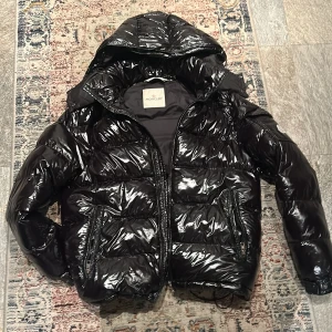 Svart pufferjacka från Moncler - Säljer en svart, glansig pufferjacka från Moncler med huva och dragkedja. Jackan har quiltad design, två sidofickor med dragkedja och coolt serietryck på insidan. Perfekt för dig som vill ha en varm och snygg jacka med streetkänsla. Pris kan diskuteras 
