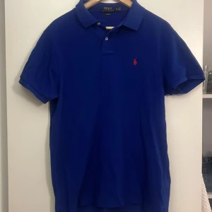 Blå pikétröja Polo Ralph Lauren XL - Snygg marinblå pikétröja från Polo Ralph Lauren i slim fit-modell. Klassisk krage, korta ärmar och två knappar framtill. Ikonisk röd logobrodering på bröstet. Perfekt för dig som gillar stilrena och sportiga plagg.