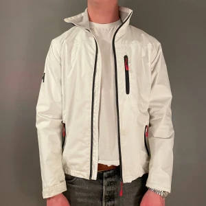 Vit vindjacka från Helly Hansen XS - Snygg vit vindjacka från Helly Hansen med svart HH-logga på ärmen och detaljer i rött vid dragkedjorna. Jackan har ståkrage, tre fickor med dragkedja och regular fit. Perfekt för dig som gillar stilren och sportig look. Jackan har några små fläckar som knappt syns. Jackan skickas 24h efter köp! Hör av dig vid minsta fråga!! (Modellen är 176 och väger 65 kg.)