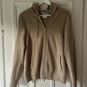 Beige hoodie i ull från Gran Sasso - Snygg beige hoodie från Gran Sasso, tillverkad i Italien. Tröjan är stickad i mjuk kashmir, har huva med snörning, dragkedja framtill och två fickor. Ribbstickade muddar vid ärmslut och nederkant ger en schysst look. Perfekt för dig som gillar stilren och enkel design.